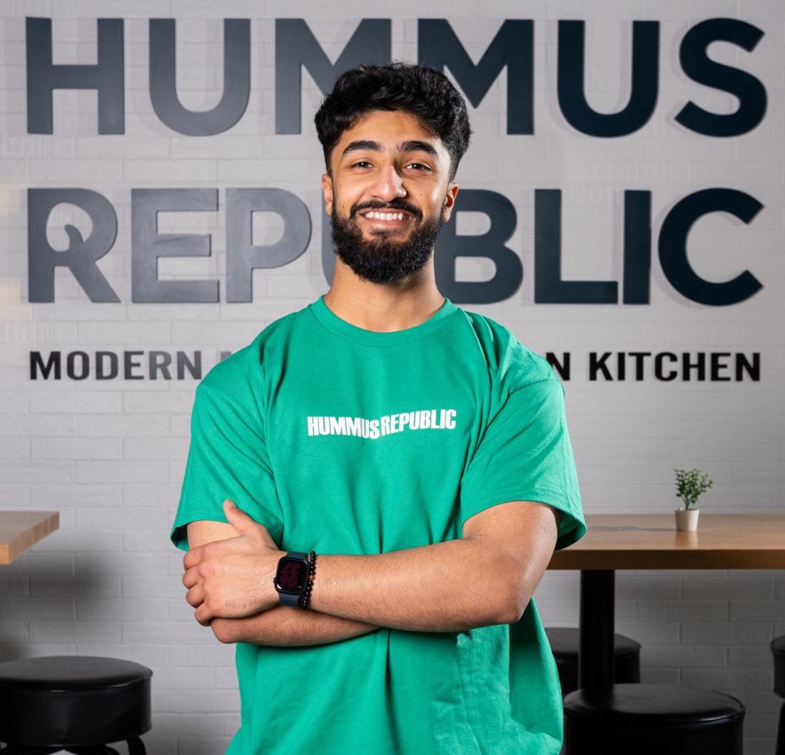Hummus Republic franchise owner standing in front of the Hummus Republic wall sign in his restaurant