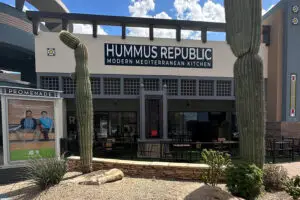 The image showcases the entrance of Hummus Republic: Modern Mediterranean Kitchen, one of the top Mediterranean restaurants near me. This sleek building features large glass windows, two tall cacti in the foreground, and a poster of two people sitting displayed on the left side.