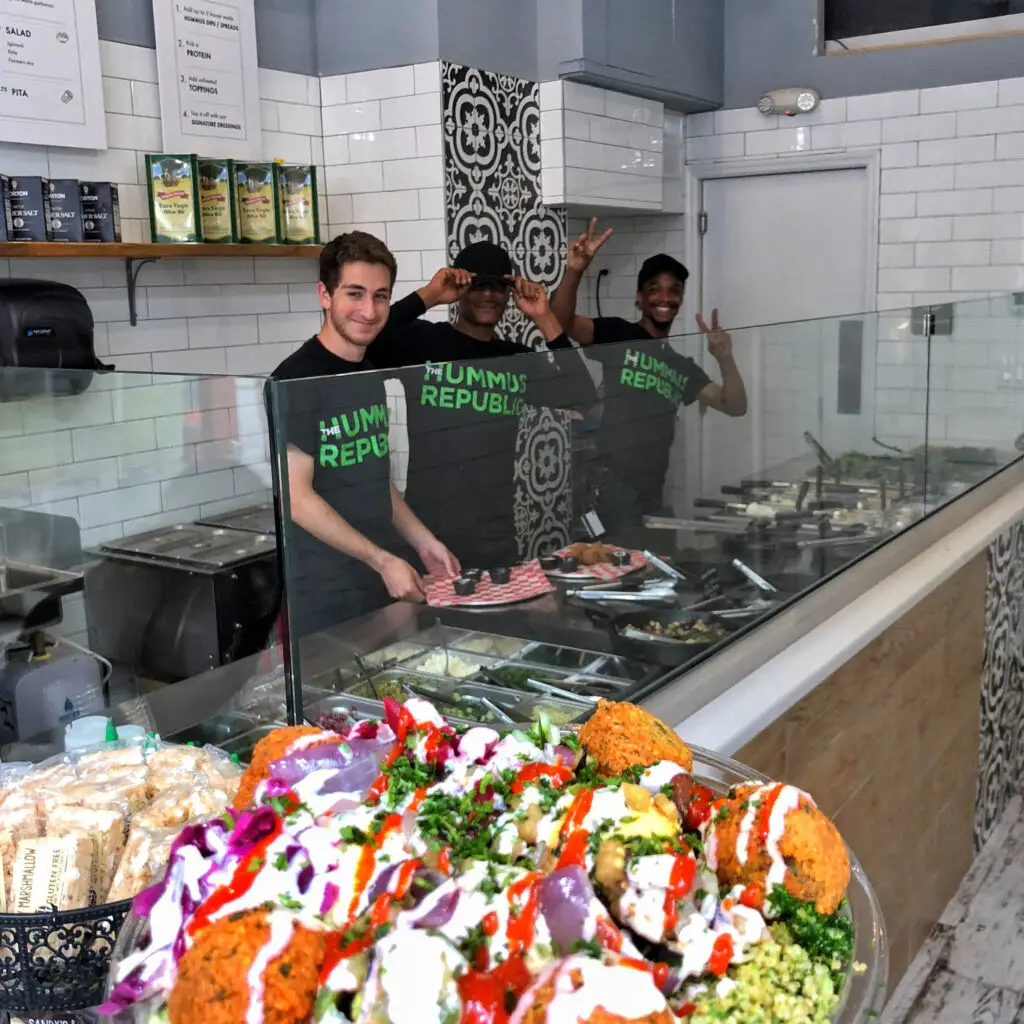 A colorful salad bowl with falafel, onions, tomatoes, and drizzled sauces is in the foreground. Behind a counter with food trays in this modern Mediterranean restaurant, three smiling staff members pose cheerfully. Two of them flash peace signs, all wearing matching Hummus Republic T-shirts.