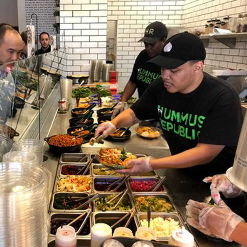 Employees at a Mediterranean restaurant serve customers a variety of fresh toppings from a counter. The workers wear black shirts with “HUMMUS REPUBLIC” in green. The setting, complete with a glass partition, bowls of ingredients, and bright white-tiled walls, creates a modern, clean atmosphere.