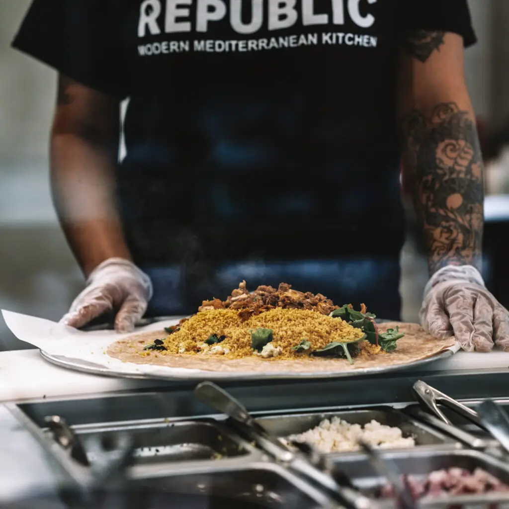 A person with tattooed arms and gloves artfully assembles a wrap at a bustling food counter. Fresh greens and grains adorn the flatbread, while the dark shirt proudly sports REPUBLIC Modern Mediterranean Kitchen. This vibrant lunch spot is a standout among restaurants near me, with an inviting array of flavors in open view.