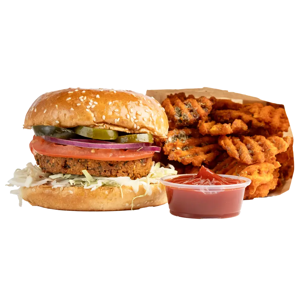 A veggie burger on a sesame seed bun, topped with fresh lettuce, tomato, red onion, and pickles, is served with waffle fries and a clear cup of ketchup. Perfect for those exploring new lunch places near me seeking delicious bites.