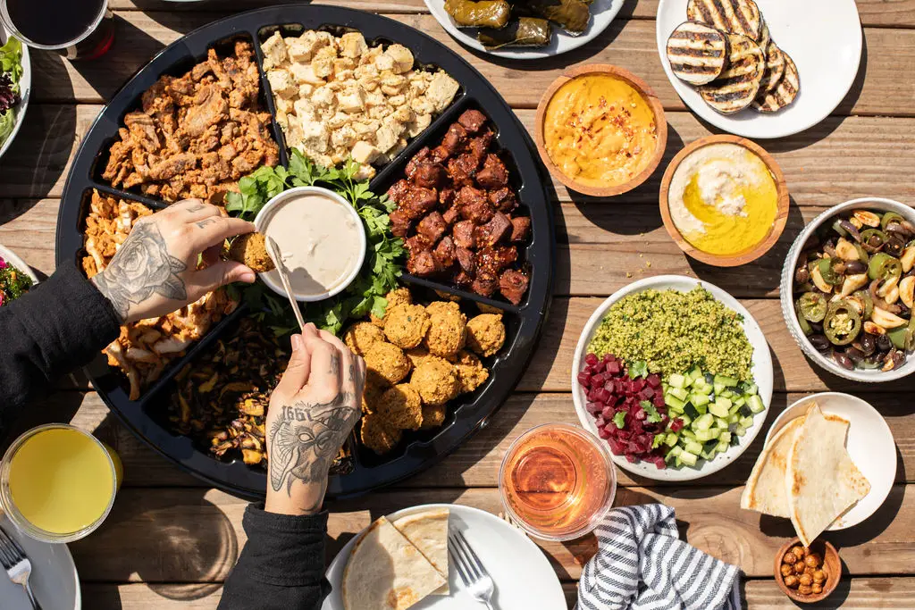 A person with tattooed hands serves dip onto a plate from a large platter of falafel, grilled meats, salads, and sauces. The table, set against an outdoor wooden background, evokes the vibrant flavors found at Mediterranean restaurants near me. Bowls of hummus and drinks accompany the meal.