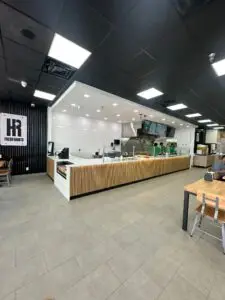 A modern restaurant interior with a sleek, minimalist design. The counter features a wooden facade and glass display. Staff members prepare food behind the counter. With tiled floors, a black ceiling, and wooden chairs, it's no wonder "Fresh Habits" is among the top Mediterranean restaurants near me.