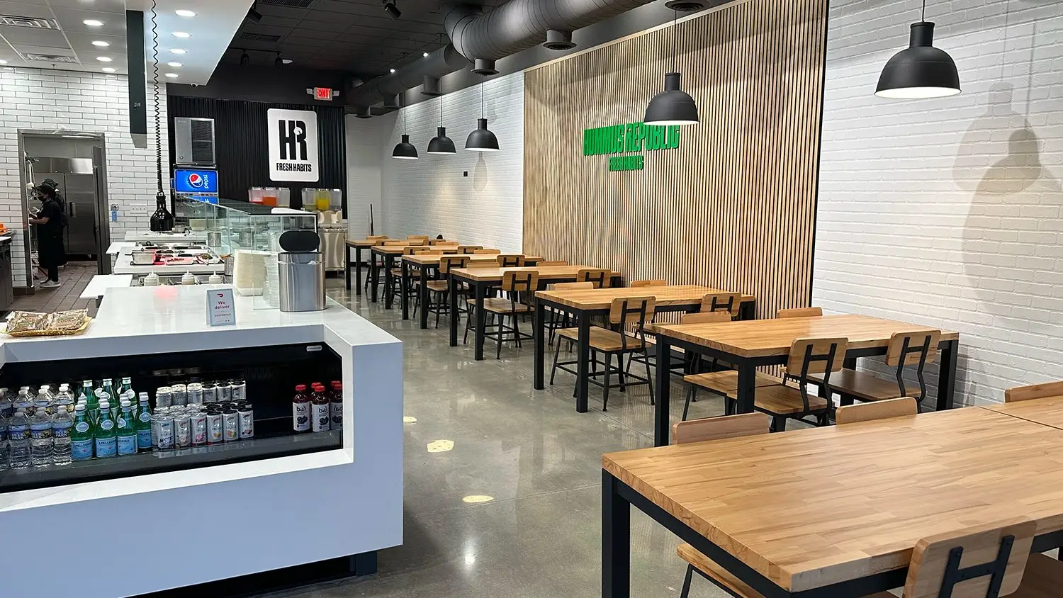 A modern Mediterranean restaurant interior features sleek wooden tables and black chairs against a white brick wall. Hanging black pendant lights illuminate the space. A counter displays beverages and a soda fountain, while the polished concrete floor reflects the minimalist design.