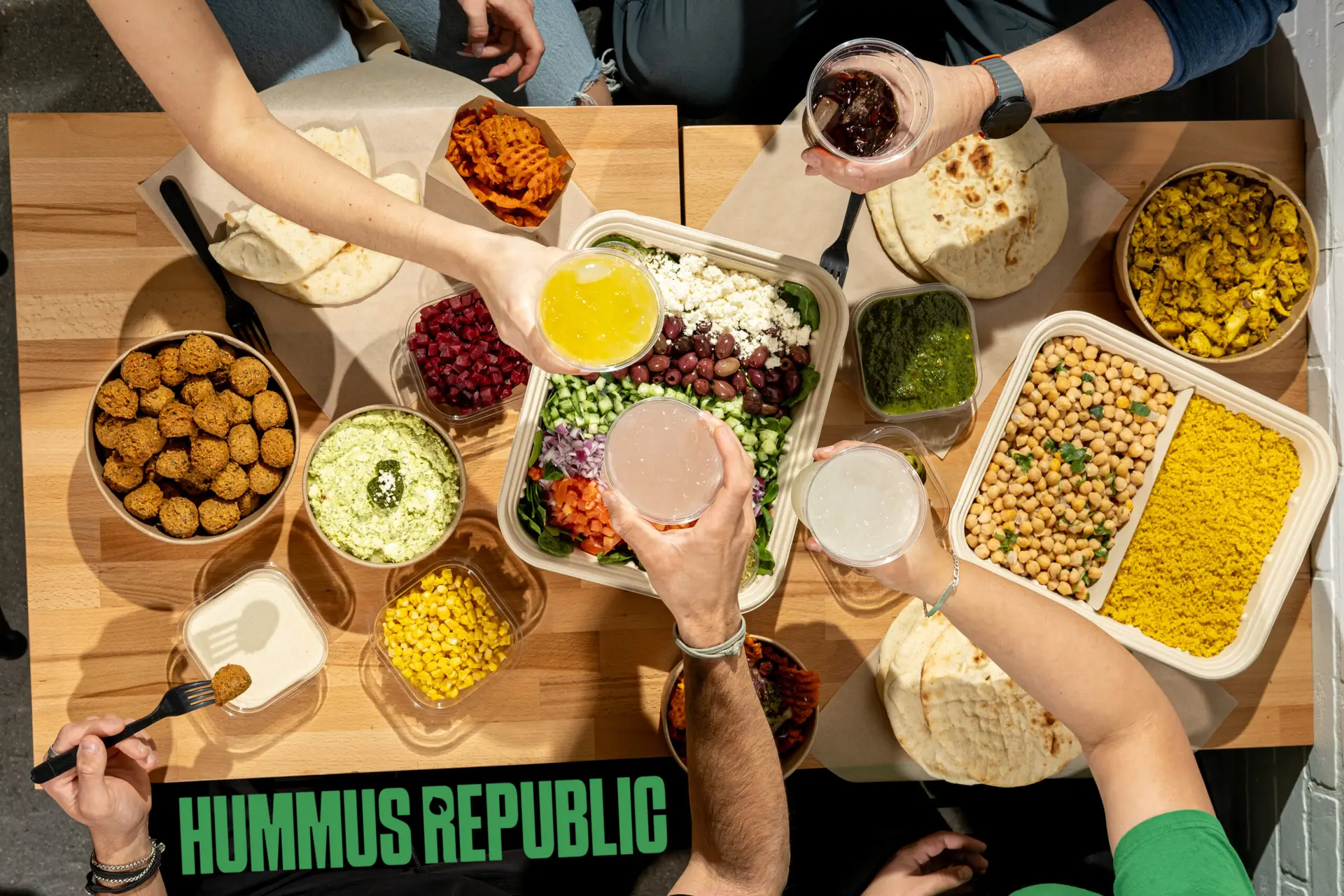 Top-down view of a table brimming with bowls of chickpeas, corn, falafel, pita, hummus, greens, and dips. Four people raise glasses filled with colorful drinks over the delicious spread at Hummus Republic—a highlight among Mediterranean restaurants near me.