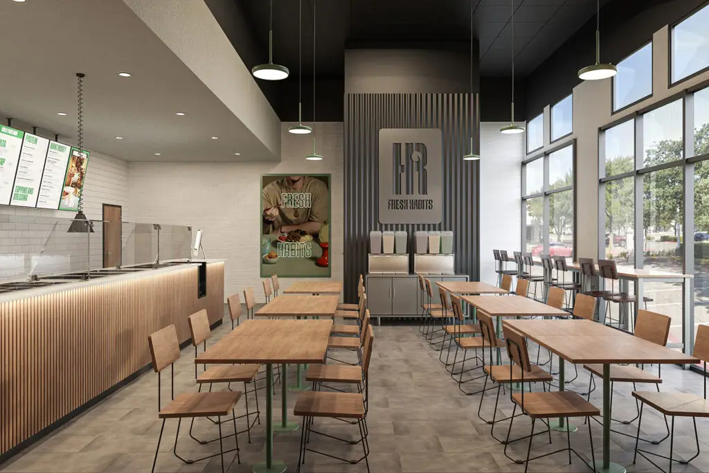 Modern restaurant interior with wooden tables and chairs, a counter on the left featuring digital menu screens, and a drink station on the right. Large windows let in natural light, while wall posters showcase appetizing imagery. Neutral tones and clean lines create a minimalist aesthetic, making it one of the most inviting lunch places near me.