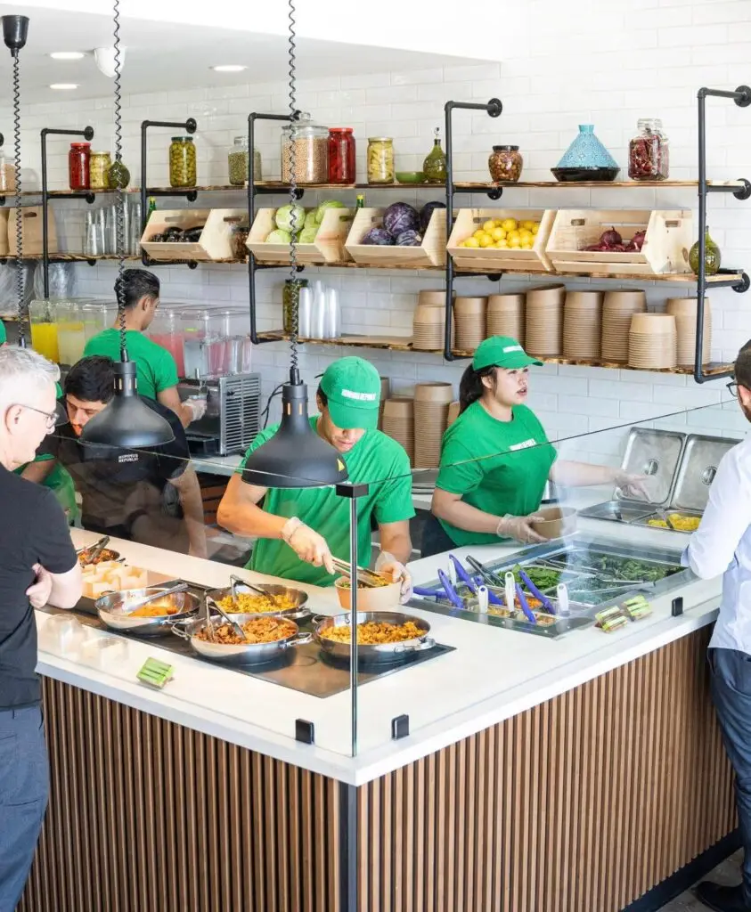 Workers in green shirts serve food at a modern deli counter with wooden accents, creating one of the best lunch places near me. Customers wait in line as shelves display jars of pickles and kitchen supplies. The counter showcases various dishes in trays, all under bright lighting and a clean ambiance.