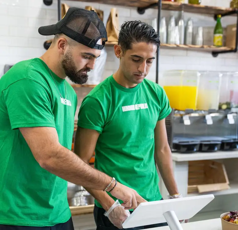 Two men wearing green T-shirts stand at a point-of-sale terminal in a Mediterranean restaurant. The man on the left points at the screen, while the man on the right observes. Shelves behind them display various jars and pitchers, including one with orange juice. Both appear focused and engaged.