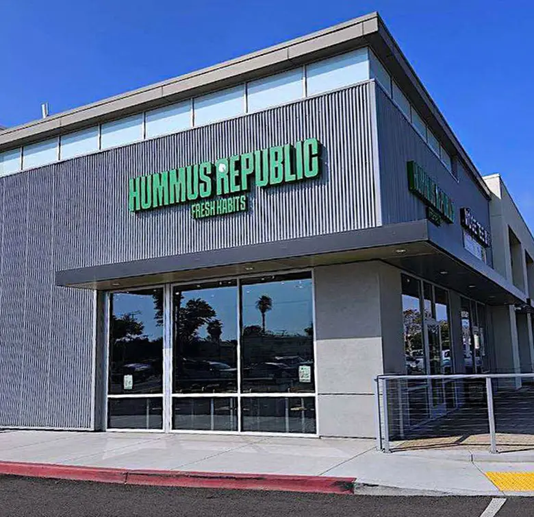 A modern building featuring the green signage of Hummus Republic Fresh Habits on a corrugated metal section, one of the best franchise opportunities. Large glass windows reflect nearby palm trees. The sidewalk is bordered by a red curb, and there's a ramp with railings leading to the entrance.