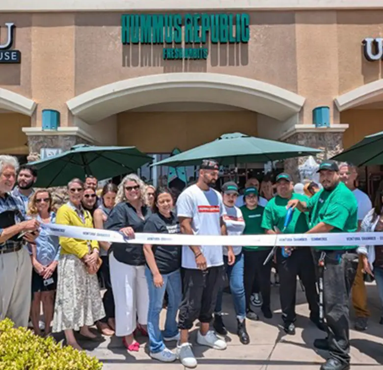 A group stands in front of a Hummus Republic restaurant for a ribbon-cutting ceremony. Holding a branded ribbon, they pose before the building's arches and outdoor umbrellas. Attendees, casually dressed, smile at the camera, celebrating one of the top Mediterranean restaurants near me.