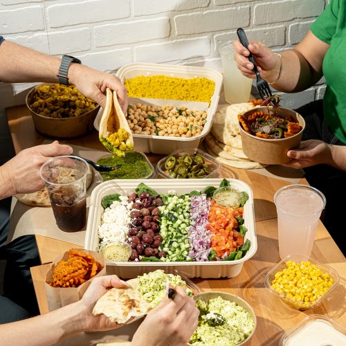 People are enjoying a meal at a wooden table in one of the best restaurants near me. Dishes include a salad with feta, olives, cucumbers, tomatoes, three curry dishes, naan, and sides of corn and sauces. Drinks feature soda and pink lemonade as hands reach in to serve and eat.