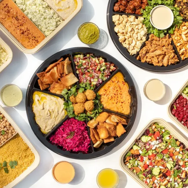 An assortment of Mediterranean dishes is displayed, including falafel, hummus, and tabbouleh, perfect for those seeking the best lunch places near me. The plates are arranged with vibrant salads featuring greens and tomatoes. Drinks and sauces are spread across a white background.